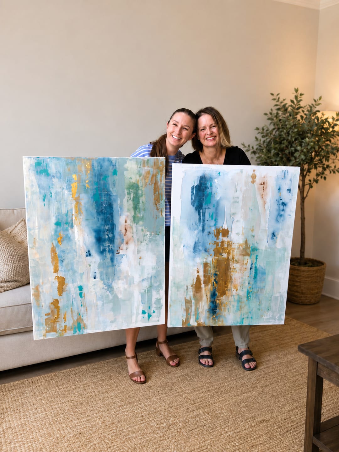 A pair of commissioned abstracts in soft blues, cream, and gold, held by two smiling clients in their living room.