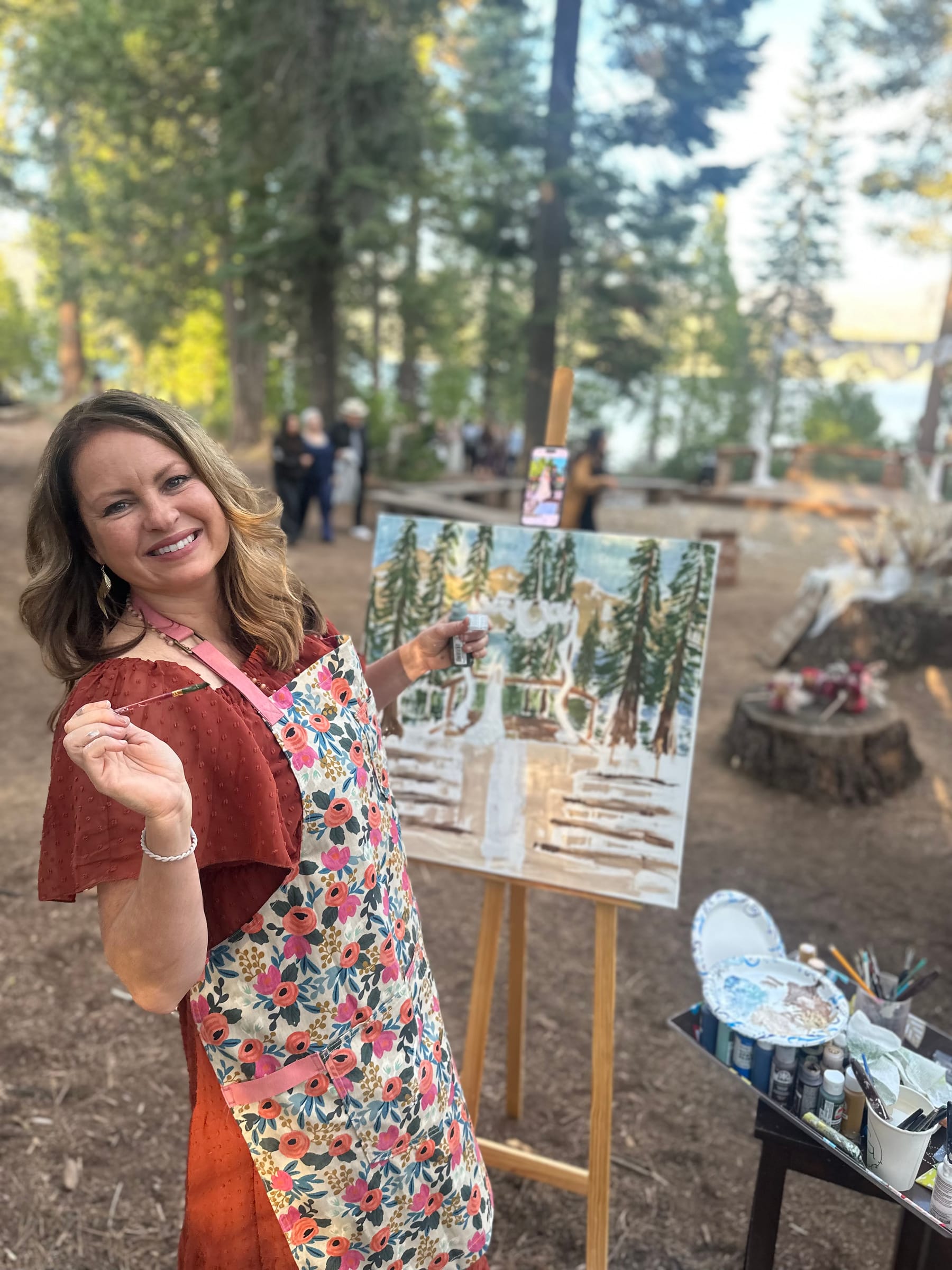 Jen Bell smiling beside her easel, brush in hand, painting the forest and lake live at an outdoor wedding at Bass Lake, California.