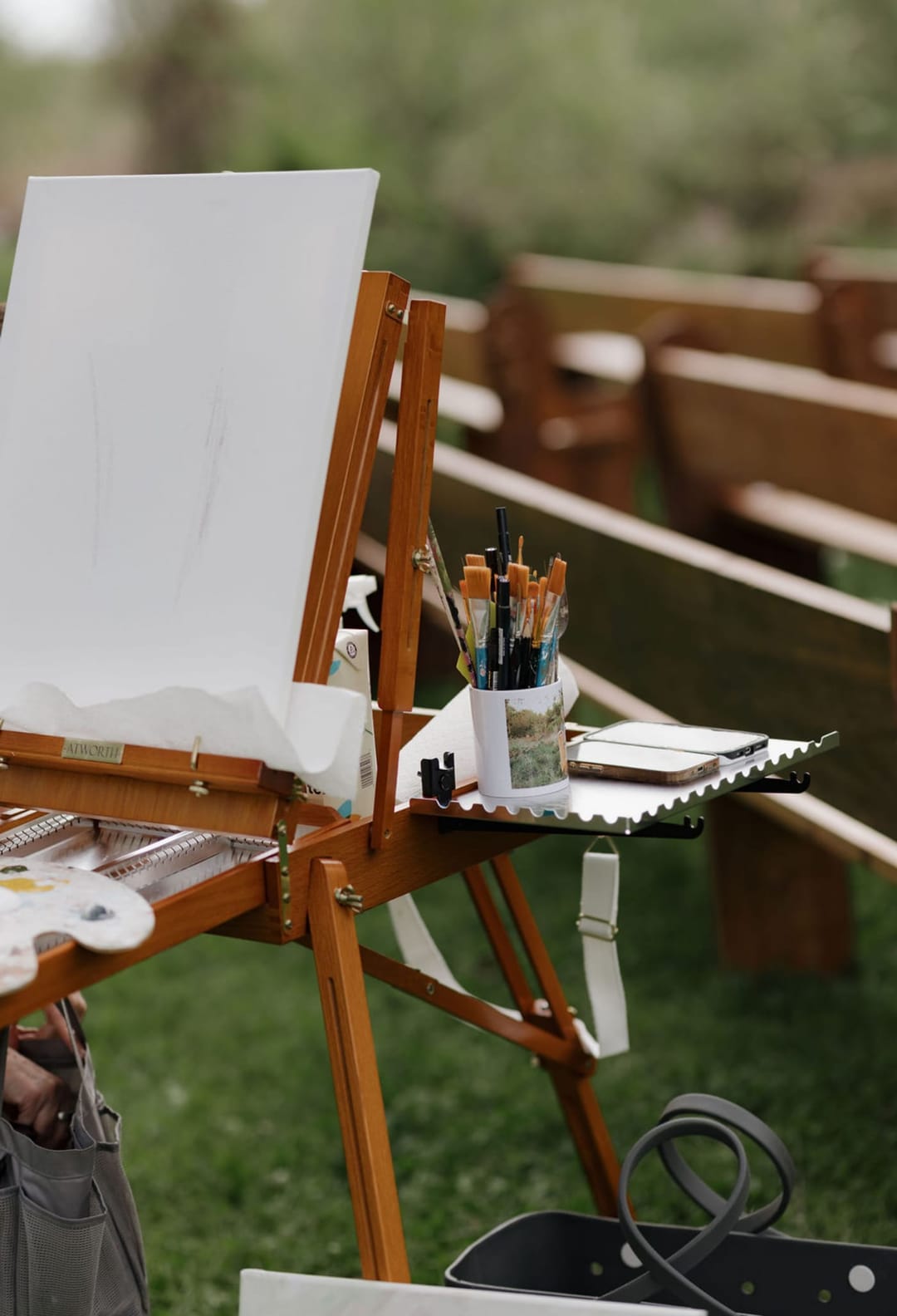 A wooden field easel holding a blank canvas, with a palette, jars of brushes, and a reference photo set up on the grass beside the wooden ceremony pews.