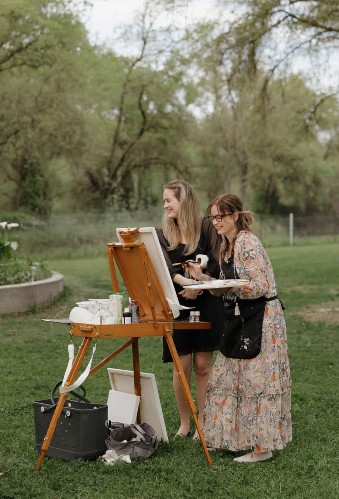 Jen standing at her easel on the lawn, palette and brush in hand, smiling as she begins to paint the scene in front of her.