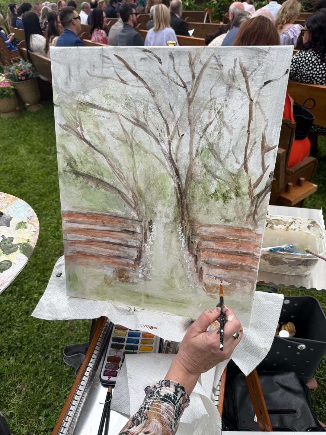 A close overhead view of the canvas mid-painting, the oak branches and tree trunks built up with detailed brushwork, Jen's brush and watercolor-style palette beside it and guests gathered behind.