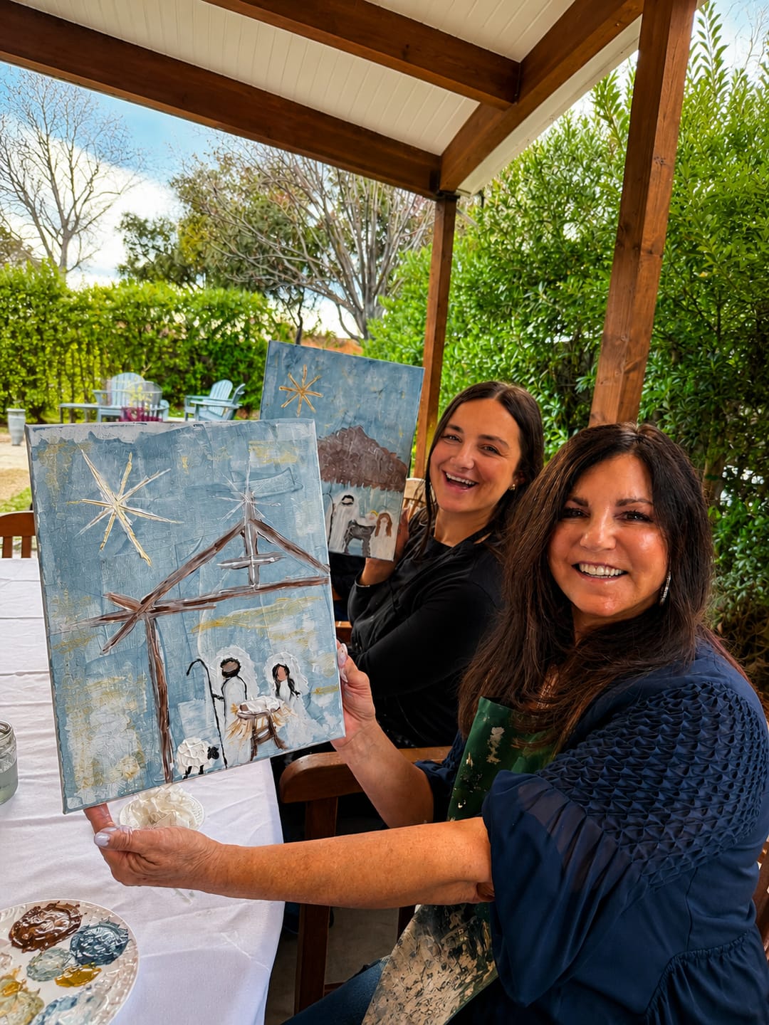Two smiling guests holding up their finished nativity paintings at an outdoor holiday workshop.